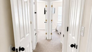 Un pasillo con puertas blancas, suelo alfombrado y vista a un dormitorio con cama y ventana.