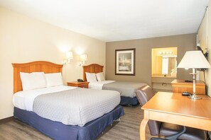 A hotel room with two beds, a desk, a chair, a lamp, a nightstand, and a framed picture on the wall.