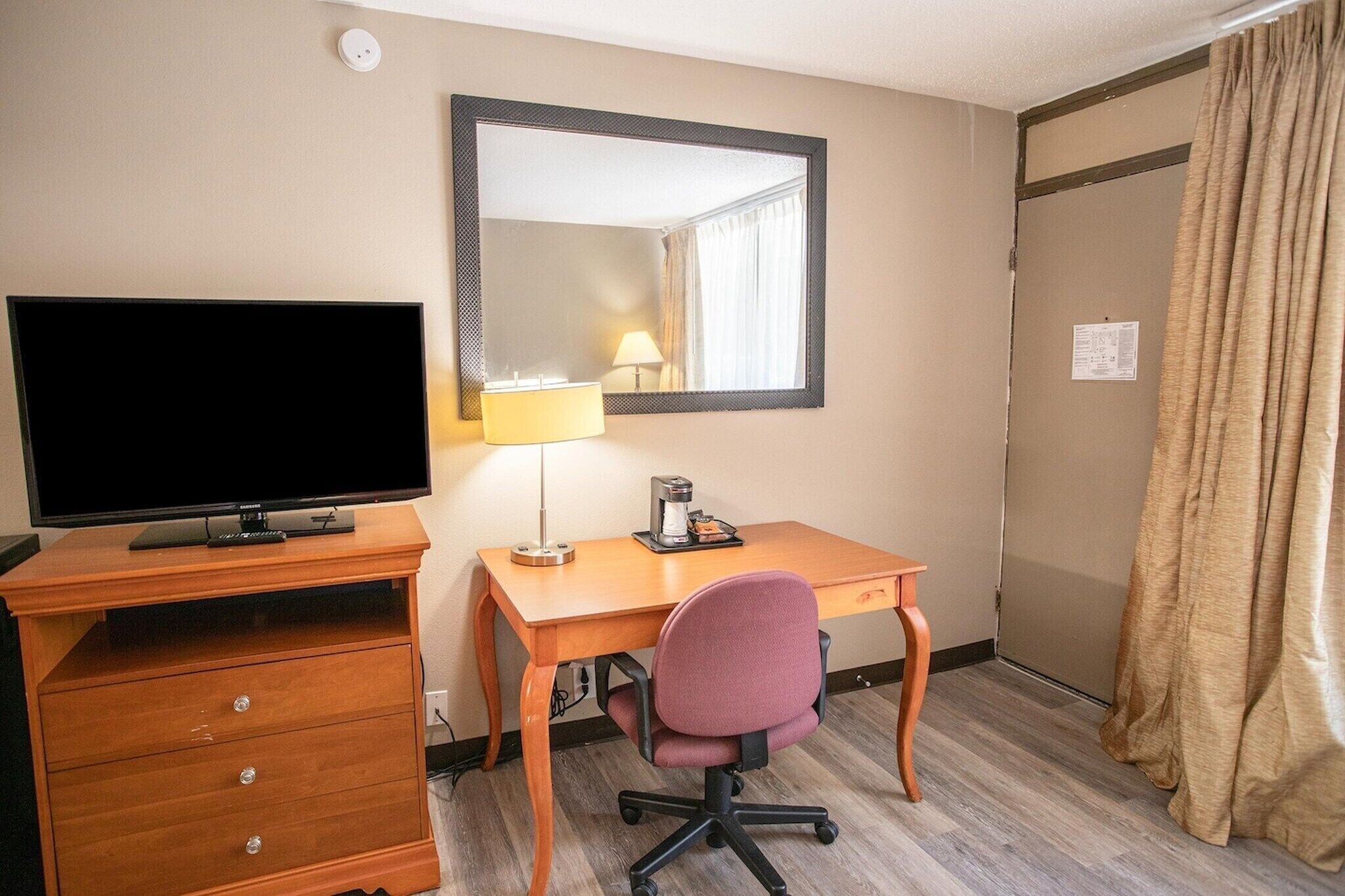 A hotel room with a wooden desk, a television, a chair, a lamp, and a mirror.