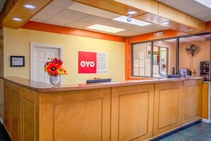 Hotellin vastaanottotiski, jossa on puinen palvelutiski, maljakko täynnä kukkia, OYO Hotels -kyltti ja seinällä oleva sertifikaatti.