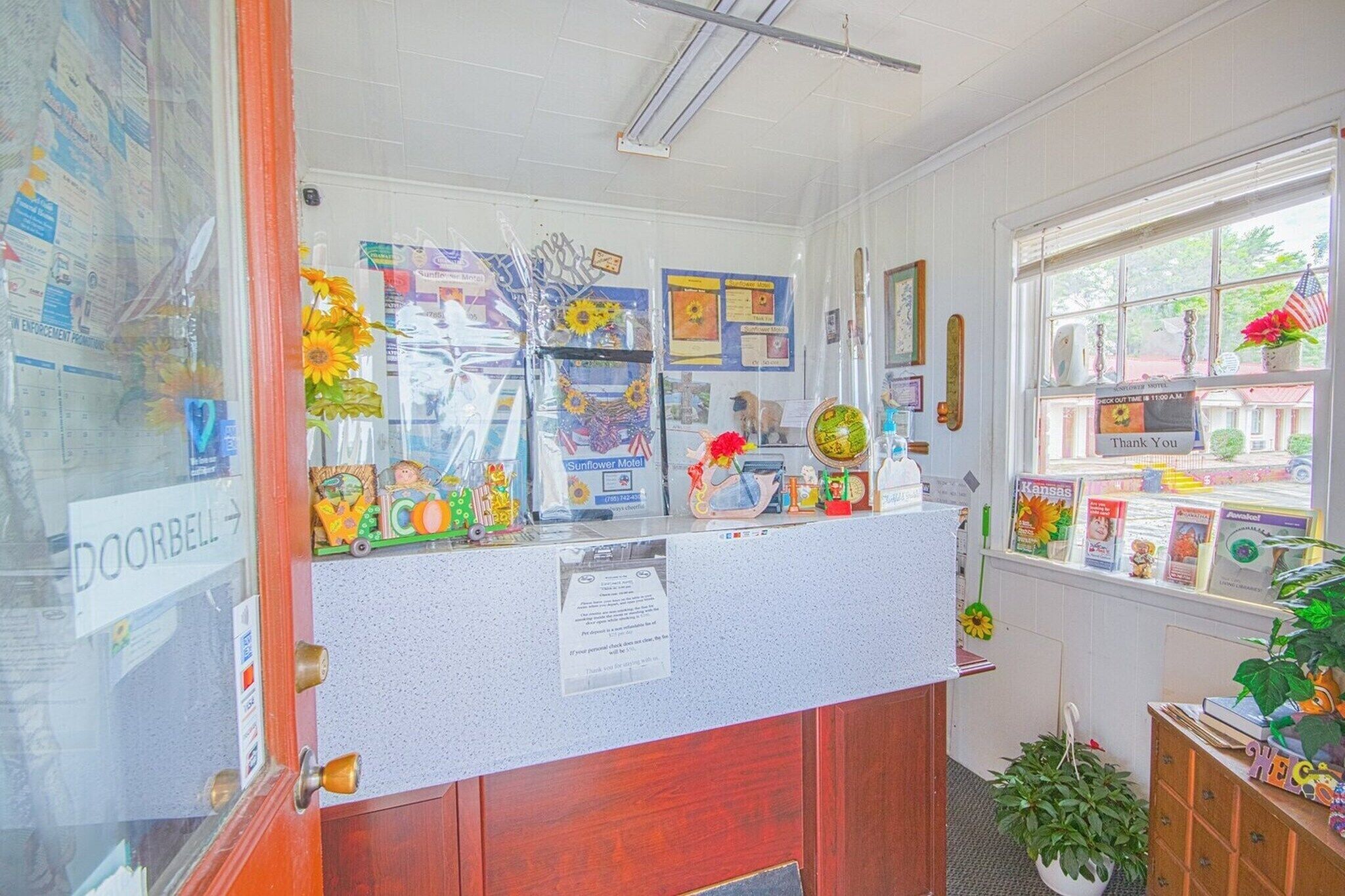 A reception desk with a 'DOORBELL' sign, a bulletin board with various notices, a window with a view of the outside, and a plant on the right.