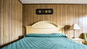 A room with a wooden paneled wall, a bed with a quilted green bedspread, a nightstand with a lamp, and a wall-mounted decoration above the bed.