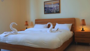 A neatly made bed with white linens, two wall-mounted lamps, and a painting on the wall. - J Apartments - 1BR @Dead Sea Resort "B24" (Dead sea hotels area next to convention center)
