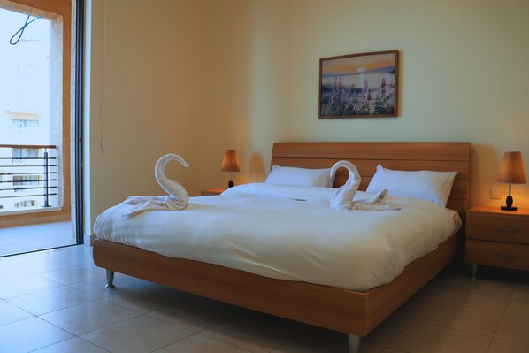 A neatly made bed with white linens and two decorative swan pillows, a wooden headboard, a nightstand with a lamp, and a painting on the wall.