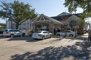 A parking lot with two cars, a building with a covered entrance, and trees. - King Bed | Non Smoking (Houston)