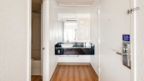 Un couloir étroit mène à une salle de bain équipée d’un lavabo, d’un miroir et d’un panneau indiquant les mesures de protection.
