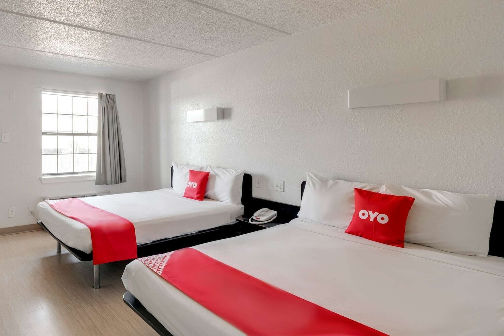 A hotel room with two beds, each with a red OYO pillow, a white wall, a window with curtains, and a white ceiling.