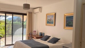 Een slaapkamer met een bed, een bureau, een stoel, een klein kastje en een groot raam.