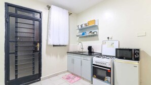 A compact kitchen with a gas stove, microwave, and refrigerator. There is a window with a white curtain, and a shelf above the sink holding kitchen items. - Cozy Studio apartment with AC in tranquil Nairobi (Nairobi)