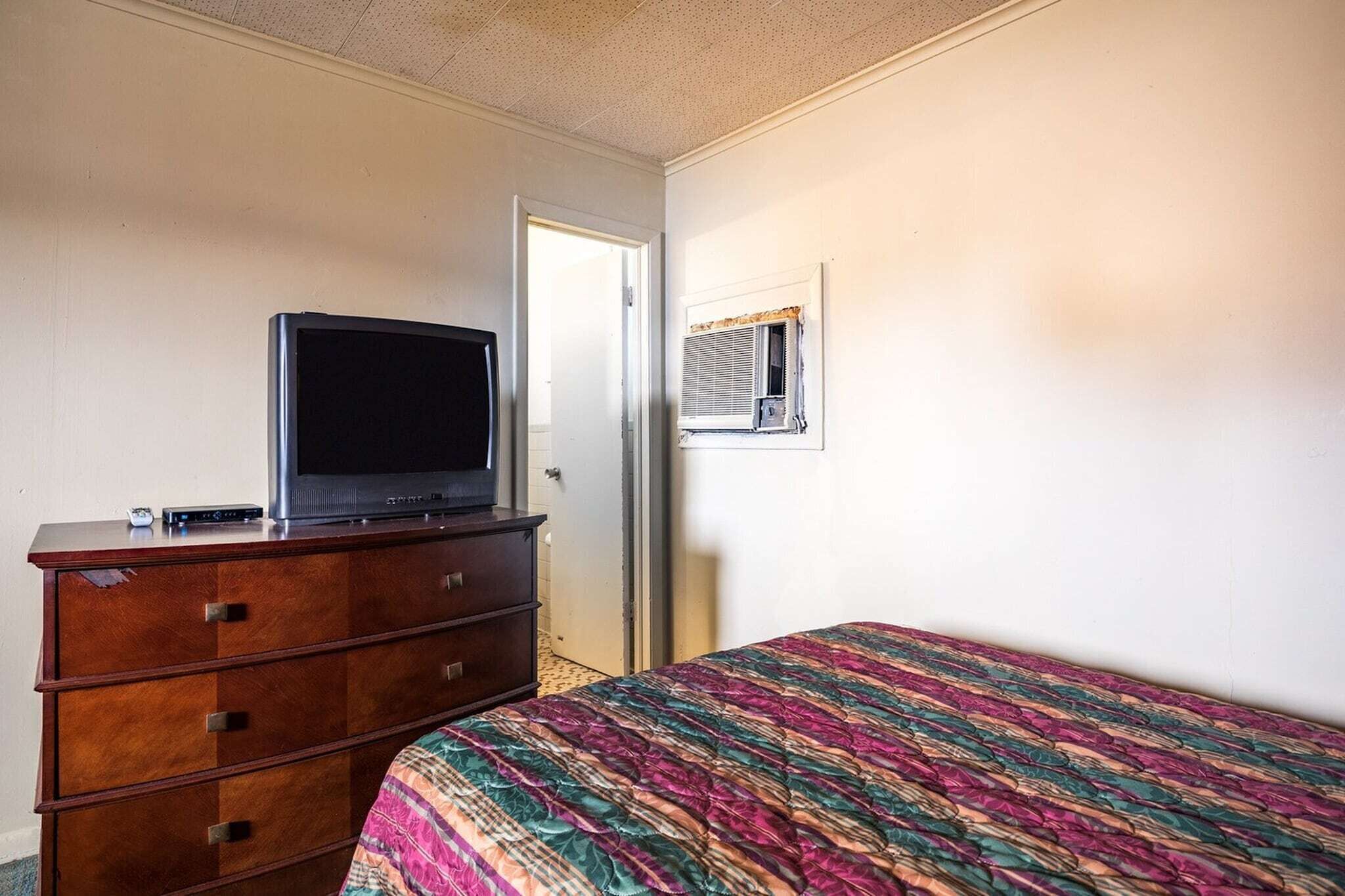 A bedroom with a bed, a dresser, a television, and an air conditioning unit.