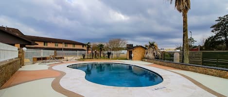 A swimming pool with a curved design, surrounded by a paved deck and a brick building with a flat roof.