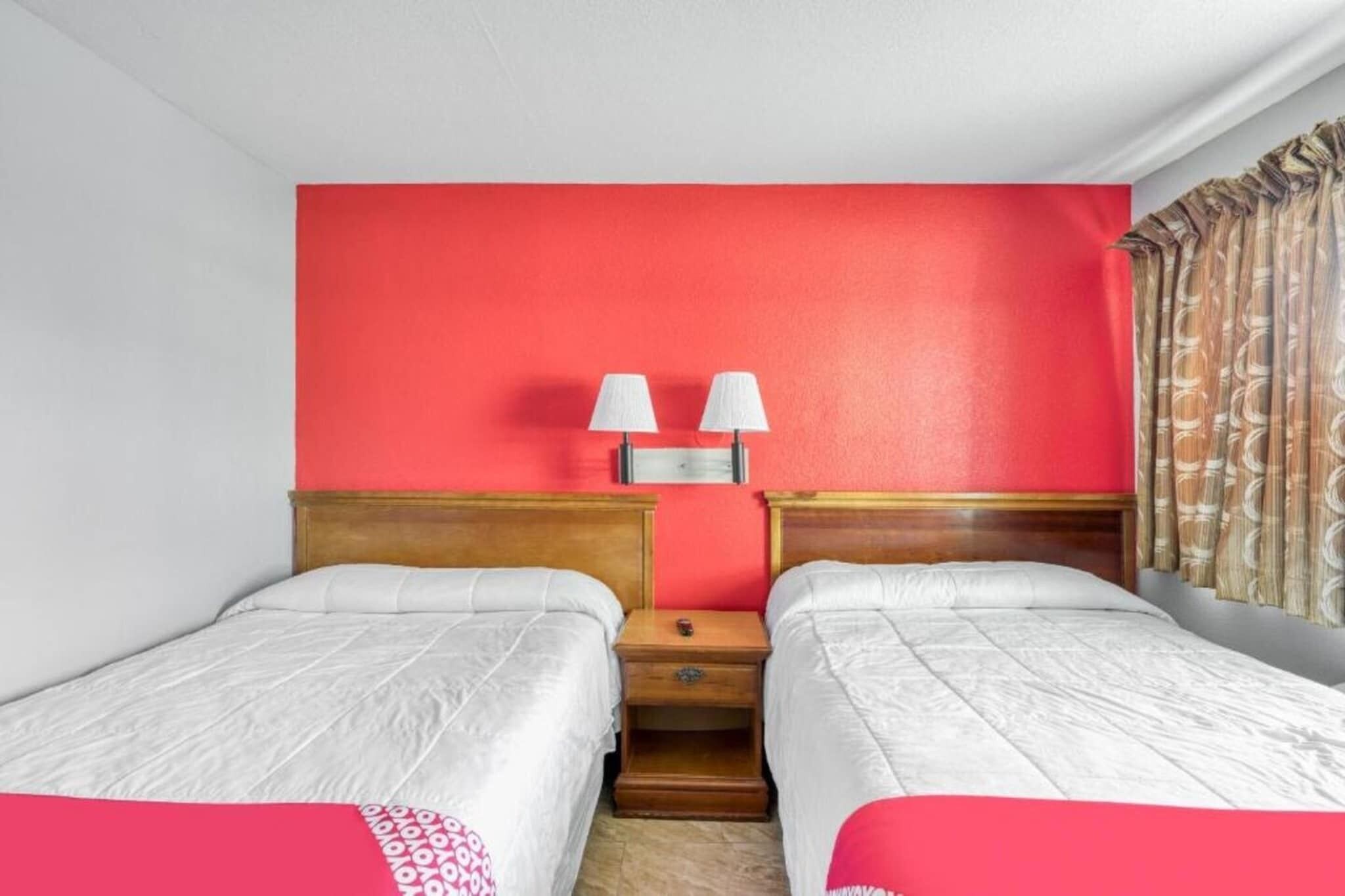 A hotel room with two single beds, a red accent wall, a nightstand with lamps, and a window with patterned curtains.