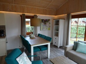 Ein Essbereich mit einem Holztisch und türkisfarbenen Stühlen, einer Couch mit Kissen sowie einem Fenster mit Blick auf Grünflächen.