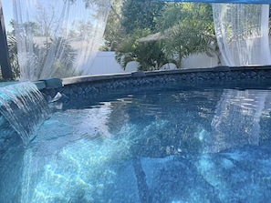 Ein klares Schwimmbecken mit Wasserfall, umgeben von einer Steinmauer und tropischen Pflanzen.