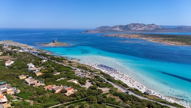 Eine Luftaufnahme einer Küstenregion mit Strand, einem Turm und einem Bergpanorama im Hintergrund.