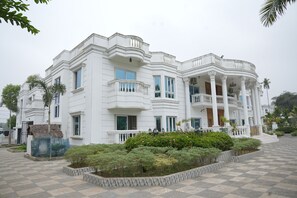 Um edifício branco grande com várias varandas e colunas, rodeado por um pavimento xadrez e vegetação.