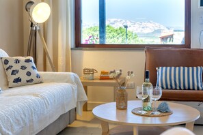 Una camera accogliente con un letto, un divano, un tavolino con bicchieri da vino e un vaso, e vista mare attraverso una finestra.