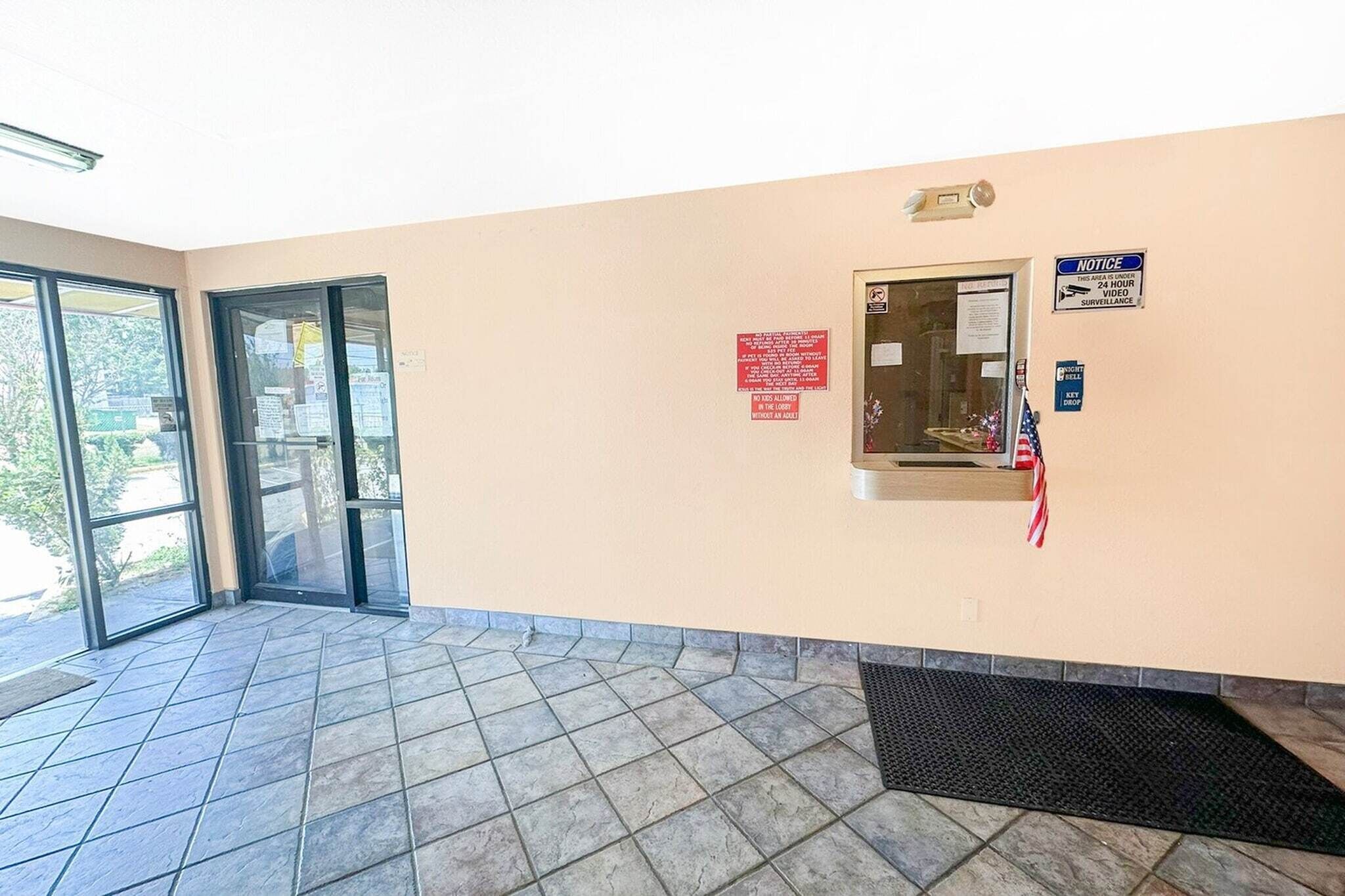 Un hall d’entrée doté d’une porte vitrée, d’un tableau d’affichage, d’un panneau indiquant la présence d’un système de surveillance et d’un paillasson à l’entrée.