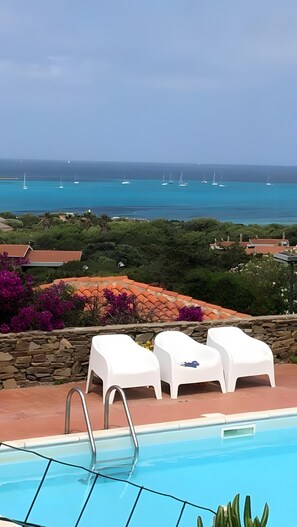 Vista della piscina con lettini, un muro in pietra e l'oceano con barche a vela.