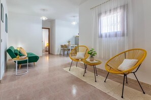 Ein modernes Wohnzimmer mit einem grünen Sofa, zwei gelben Korbstühlen, einem kleinen runden Tisch und einem Essbereich im Hintergrund.