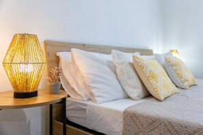 En seng med hvide og mønstrede puder, en sengetavlelampe og en lille potteplante på et trænatbord.
