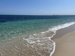 Ein Strand mit klarem Wasser, sandigem Ufer und weiter Küste im Hintergrund.