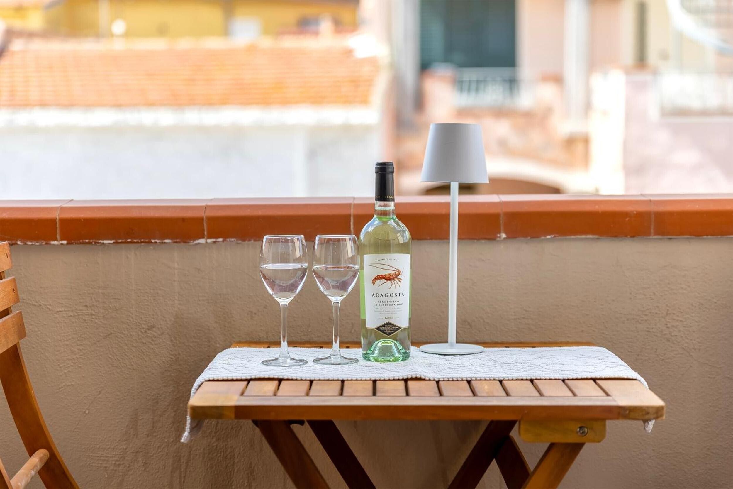 A wooden table with a bottle of Aragonese wine, two glasses, and a lamp on a balcony with a view of buildings.