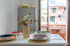 Taula de dinar posta amb plats, cuplets i gots, un gerro de vímet amb flors, i vista d'un balcó amb taula i cadires.