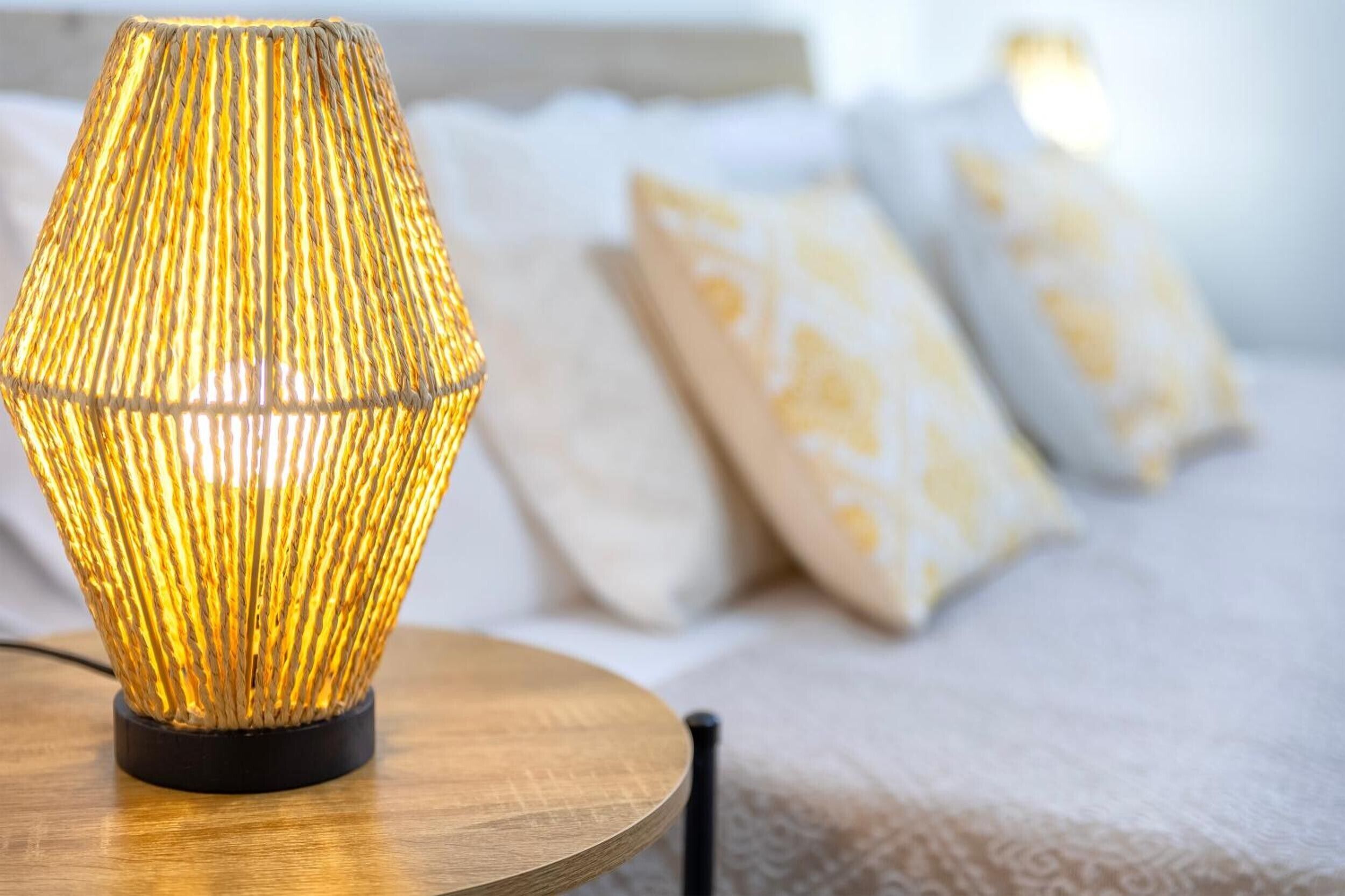 A lamp with a ribbed design on a wooden table, a bed with patterned pillows, and a white wall in the background.
