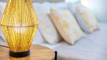 A lamp with a ribbed design on a wooden table, a bed with patterned pillows, and a white wall in the background.