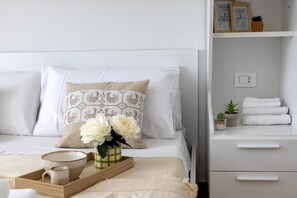Ein ordentlich eingerichtetes Hotelzimmer mit einem Bett, einem Nachttisch mit einer Auflage, einem kleinen Topfpflanzen und einem Regal mit gerahmten Bildern und einer Pflanze.