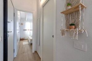 Käytävän lattia on puuta, siellä on ovi, seinälle kiinnitetty makrame-hylly, jossa on ruukkukasveja, sekä valokatkaisin.