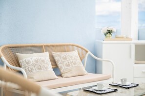 Een rieten bank met gekleurde kussens, een witte tafel met kop en schotel, en een uitzicht op de lucht door een raam.