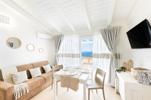 Ein Zimmer mit weißer Decke, einem braunen Sofa, einem Esstisch mit Tellern und Gläsern und Meerblick durch Vorhänge.