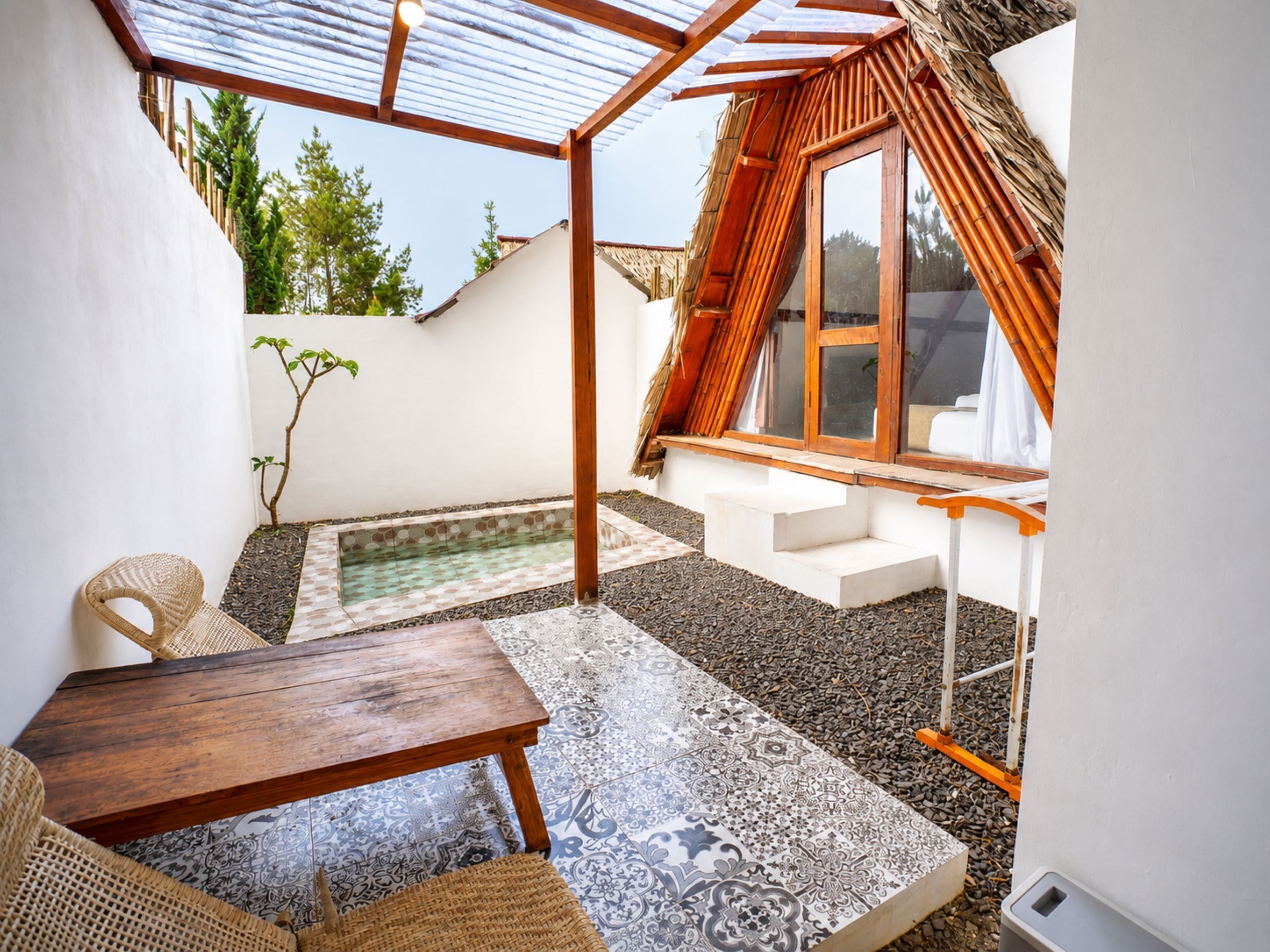 Un pequeño patio al aire libre con una mesa de madera, una silla tejida, una piscina pequeña y una estructura de techo de paja.