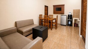 Un espacio habitable compacto con sofá, mesa de comedor con sillas, refrigerador pequeño y televisión montada en la pared.