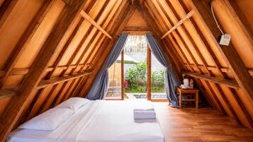 A room with a sloped ceiling, wooden beams, a bed with white linens, a small table with a kettle, and a view of greenery outside.