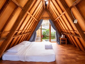 Um quarto com teto inclinado, vigas de madeira, uma cama com roupa de cama branca, uma pequena mesa com uma chaleira e vista para o verde lá fora.