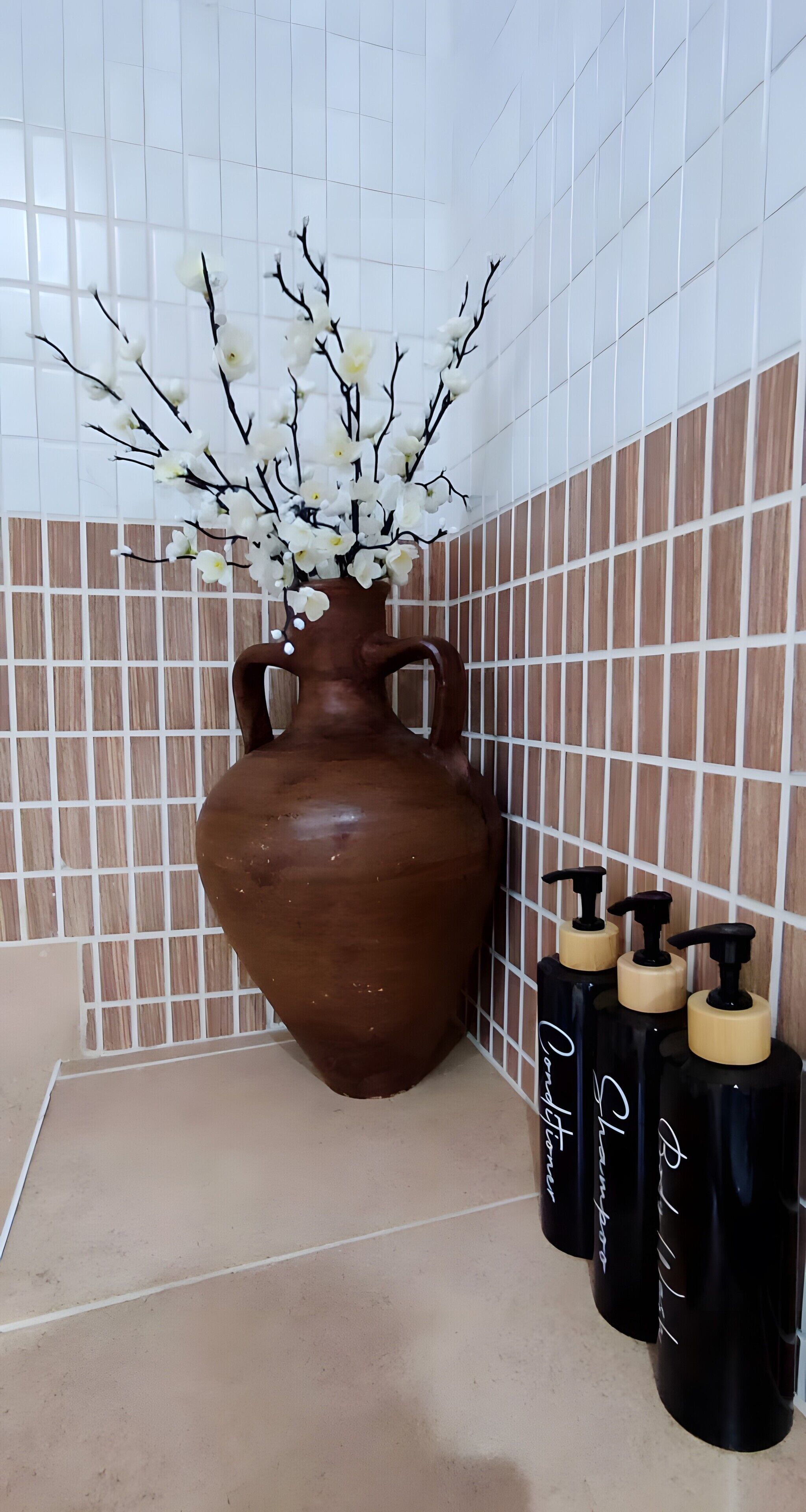 Une salle de bain avec un grand vase en céramique contenant des fleurs blanches, trois flacons de shampoing noirs et des murs recouverts de carreaux.