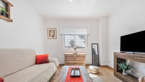 Un salon comprenant un canapé, une table basse en bois, un téléviseur posé sur un meuble et une fenêtre avec des stores.