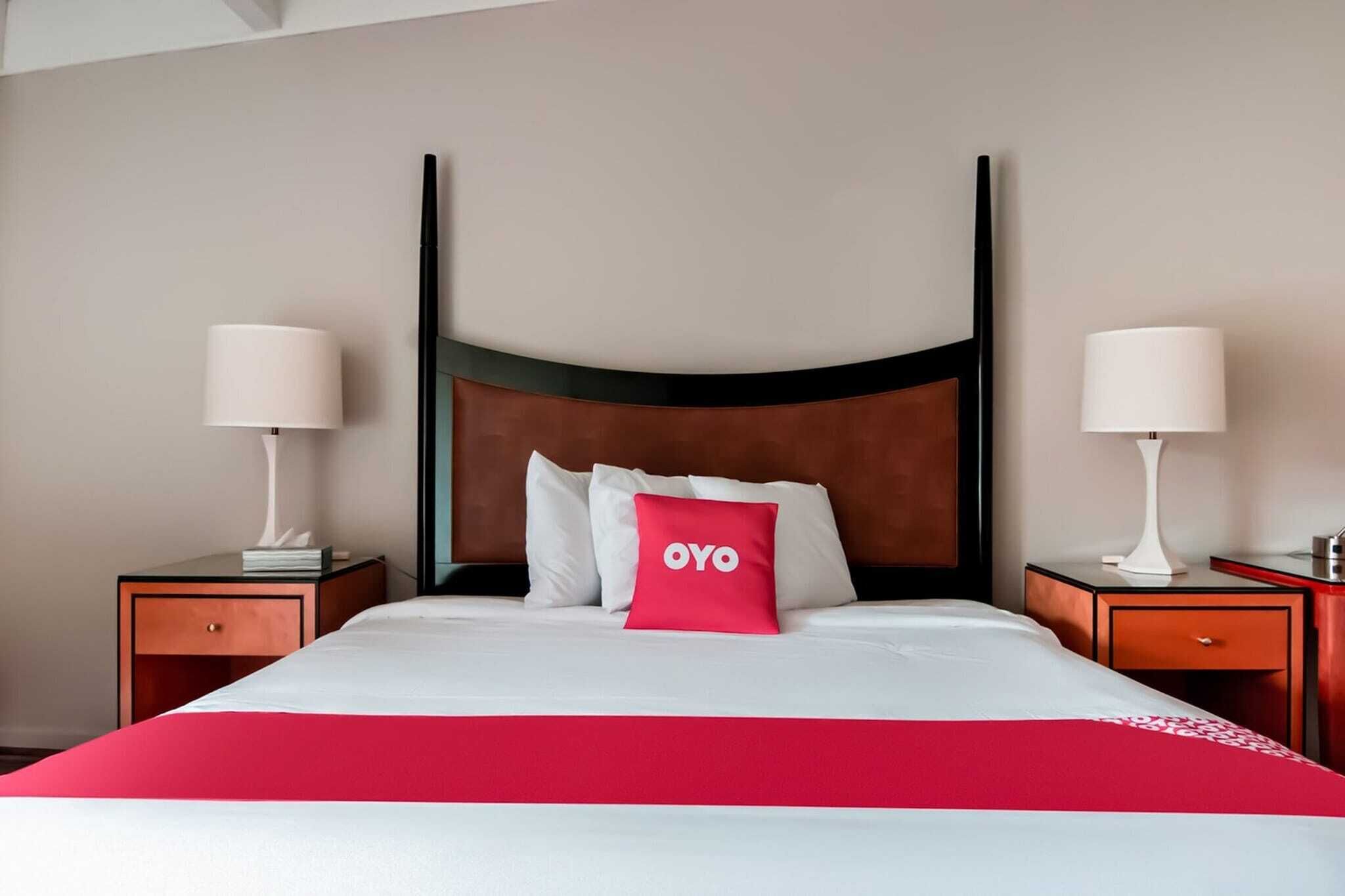 A hotel room with a four-poster bed, two bedside tables with lamps, a red throw blanket, and a pillow with 'OYO' branding.