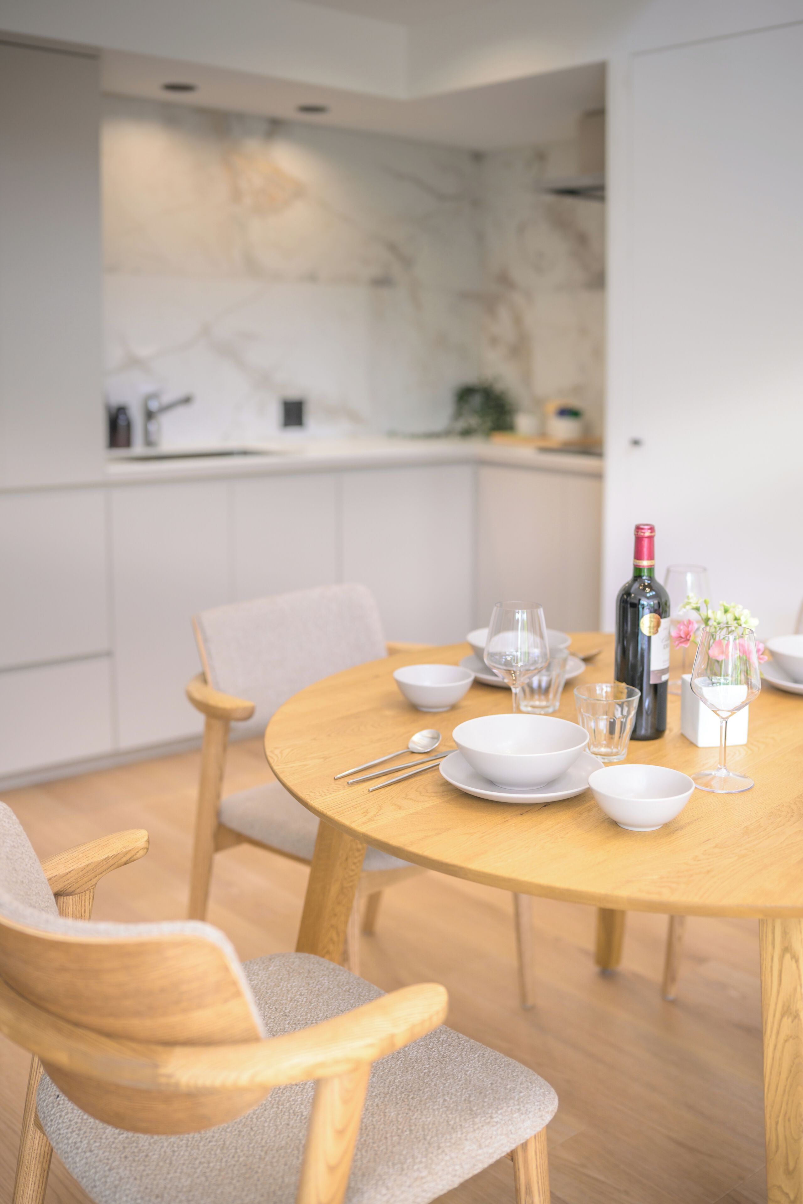 Un'area pranzo moderna con un tavolo rotondo in legno, sedie bianche e una cucina sullo sfondo.