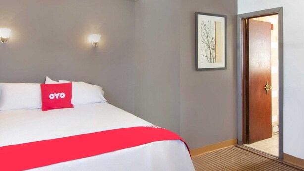 Habitación de hotel con una cama, un cojín rojo, un cuadro en la pared y una puerta a la derecha.