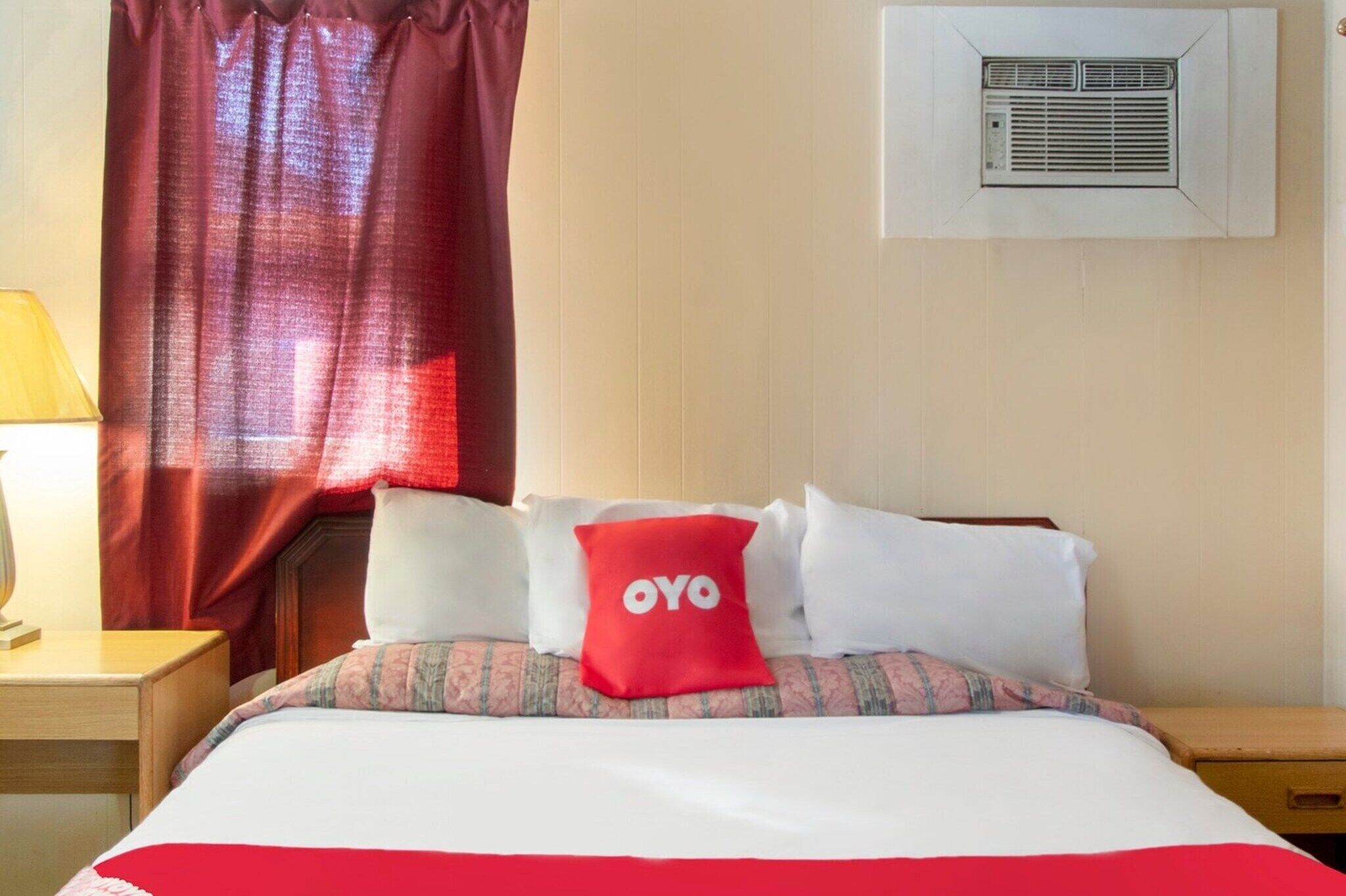 Een hotelkamer met een bed, nachtkastje, lamp, raam met gordijnen en een airconditioner.