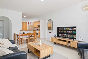 Ein modernes Wohnzimmer mit einem Holztisch, einer schwarzen Ledersofa und einem Fernseher mit digitalem Bildschirm, sowie einer Küche im Hintergrund.
