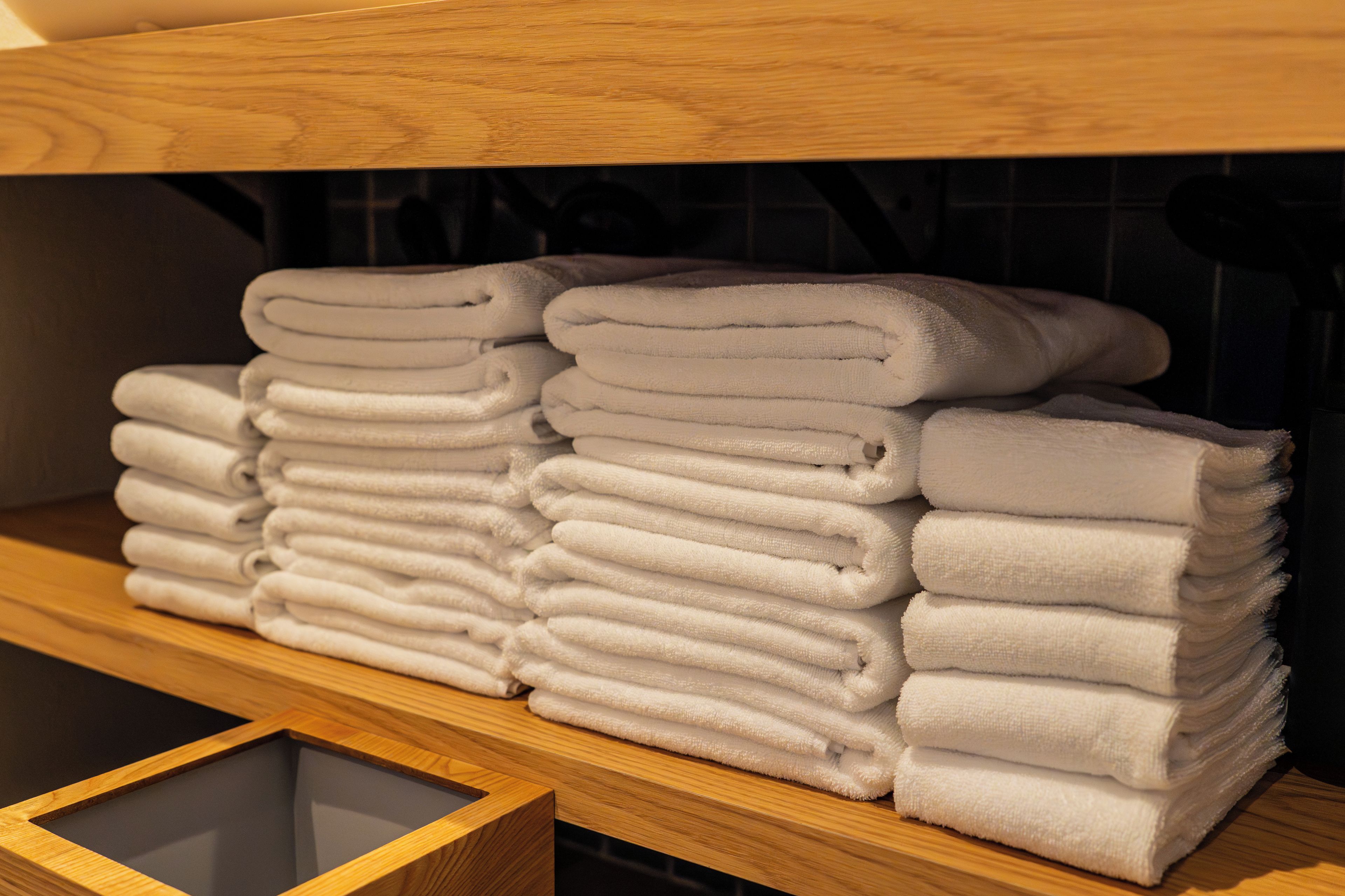 A stack of white towels on a wooden shelf in a room with a tiled wall.
