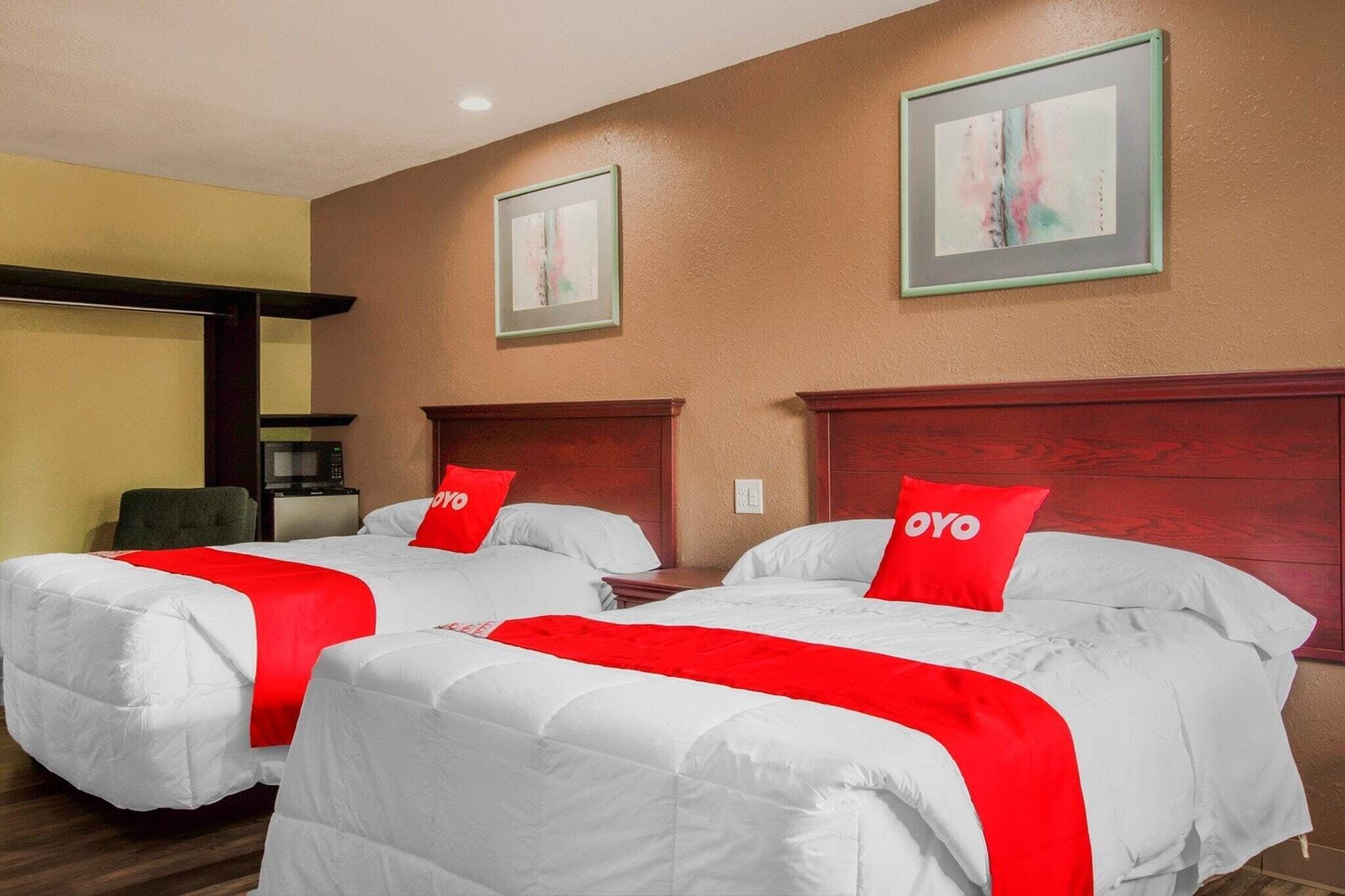 A hotel room with two beds, each with a red pillow and a white bedspread. There are framed pictures on the wall and a wooden headboard.