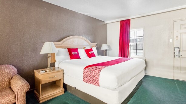 A hotel room with a bed, nightstand, lamp, chair, and a window with curtains. - King Bed|Non-Smoking (Bald Knob AR)