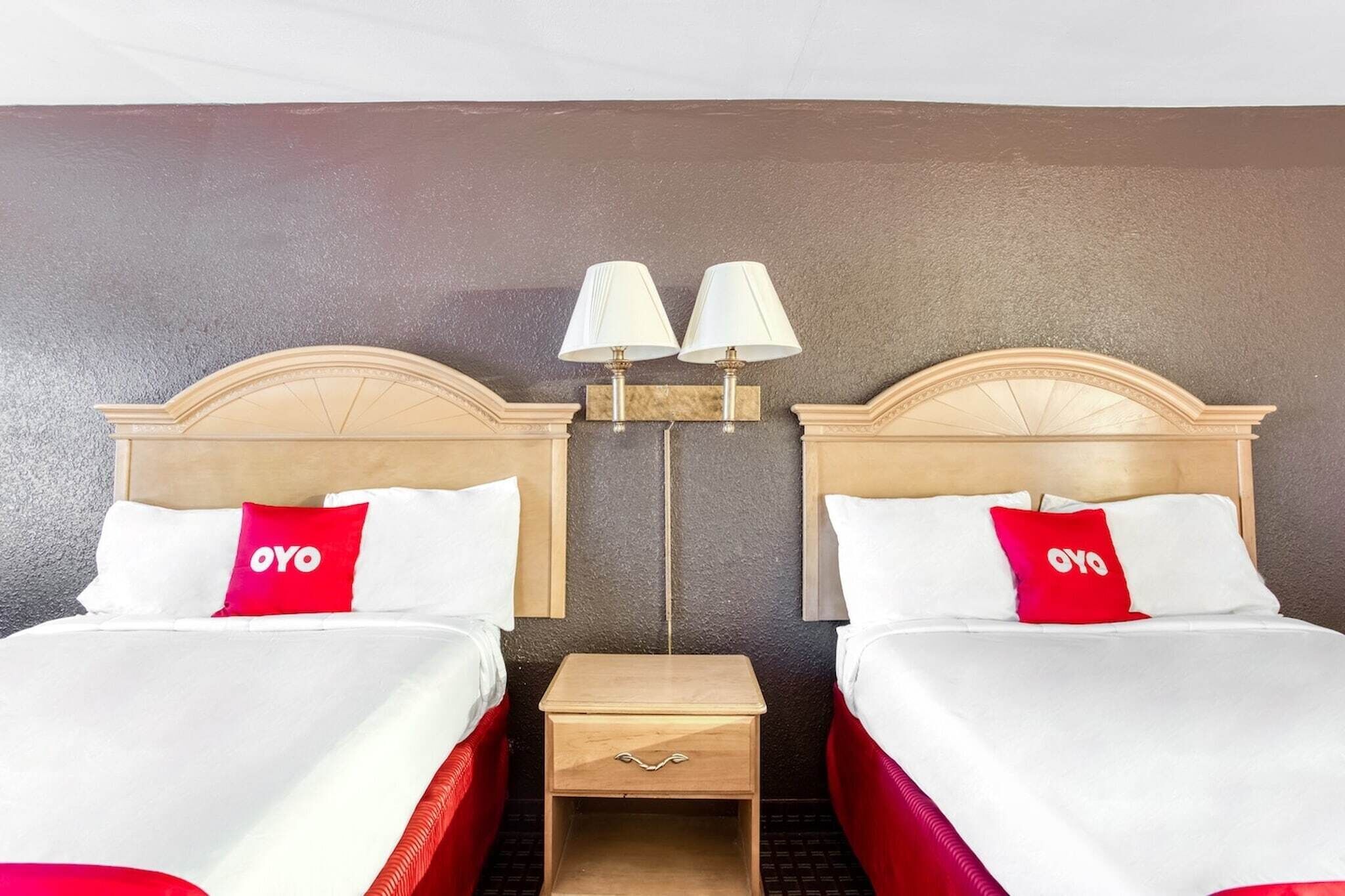 Habitació d'hotel amb dos llits, cada un amb un capçal de fusta, roba de llit blanca i almoines vermelles amb el logotip 'OYO'. Hi ha dues làmpades subjectes a la paret entre els llits.
