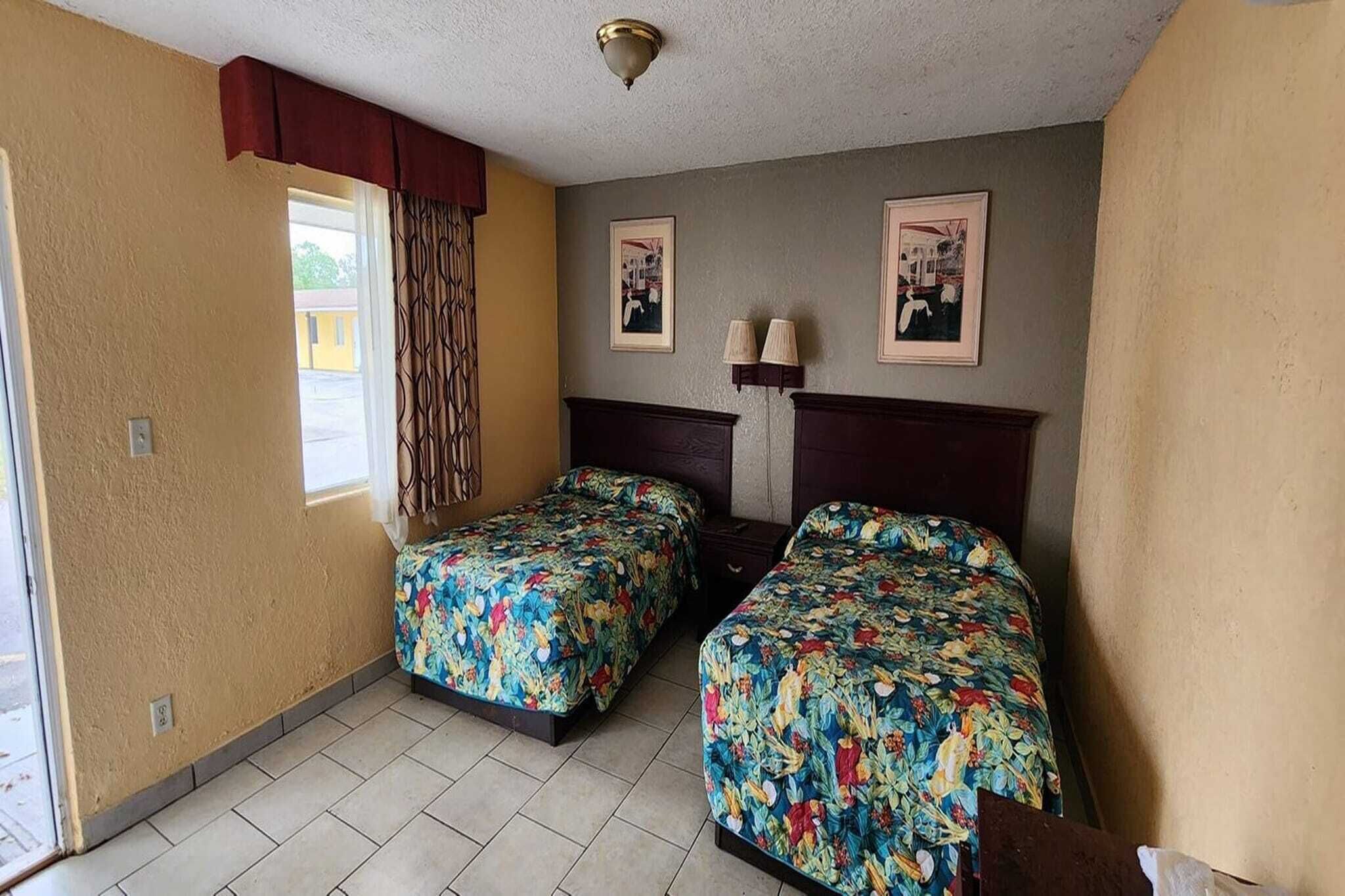 A room with two beds, a window with curtains, a wall-mounted lamp, and framed pictures.